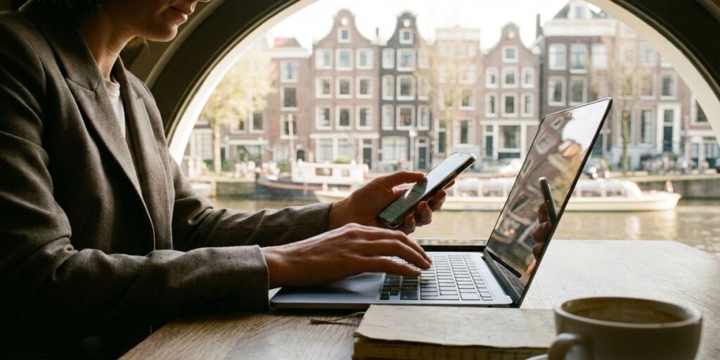 Zakenvrouw werkt op laptop en smartphone aan café tafel in Amsterdam met grachtenpanden op achtergrond